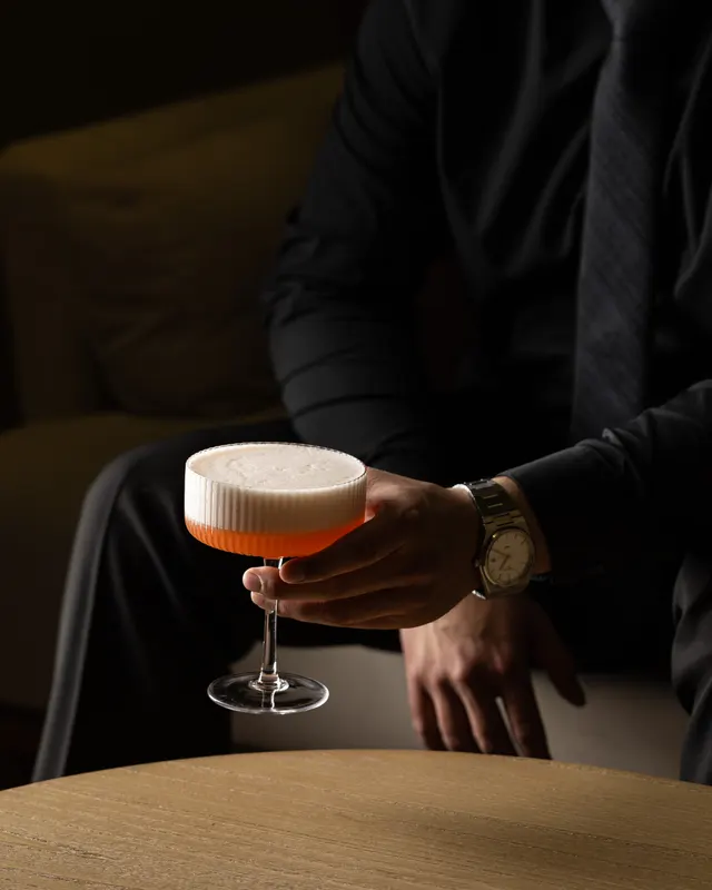 A suited guest holds a foam-topped coupe over a round oak table, shot for Barworks, Campbell