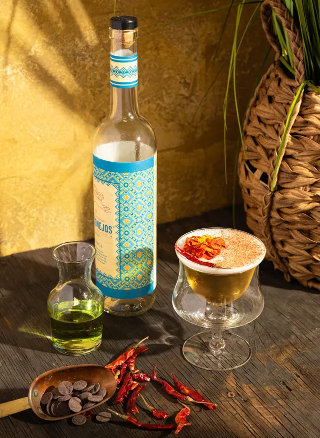 An agave bottle, a spiced coupe crowned with a marigold, and dried chiles on the wood table against a yellow wall, from Caribbean Cocktails for Ten Speed Press