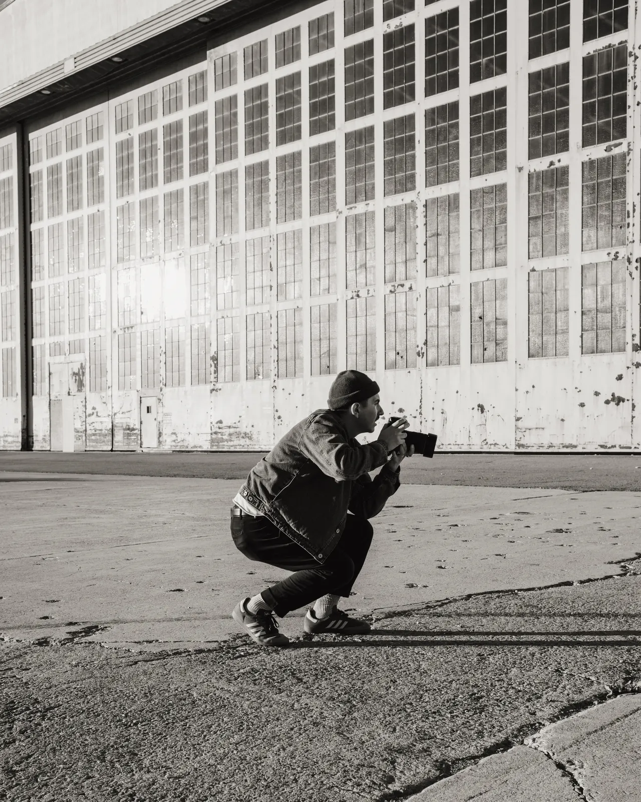 Eddie crouched low with his camera up, shooting in front of a hangar wall of windows, black and white