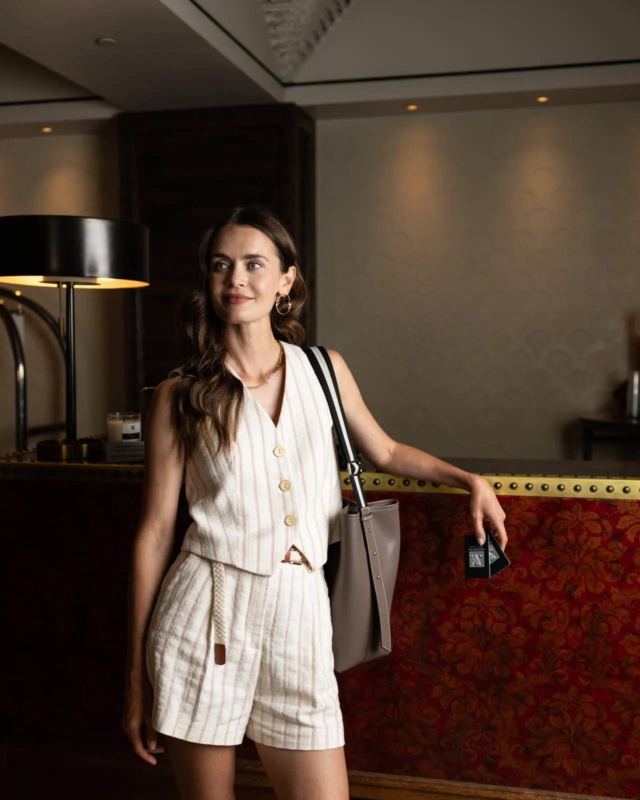 A guest in a striped linen set leans on the red damask reception desk with her key cards and tote at Hotel Valencia, Santana Row, San Jose
