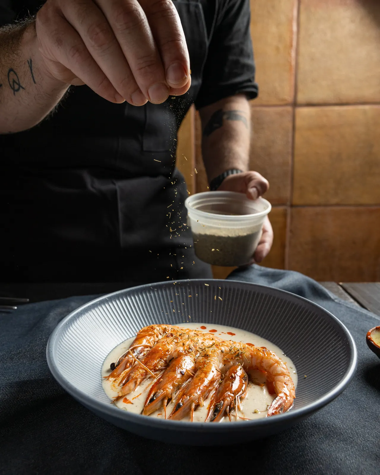 The chef sprinkling seasoning from height over the gambas in a ribbed blue bowl at La Marcha, Berkeley