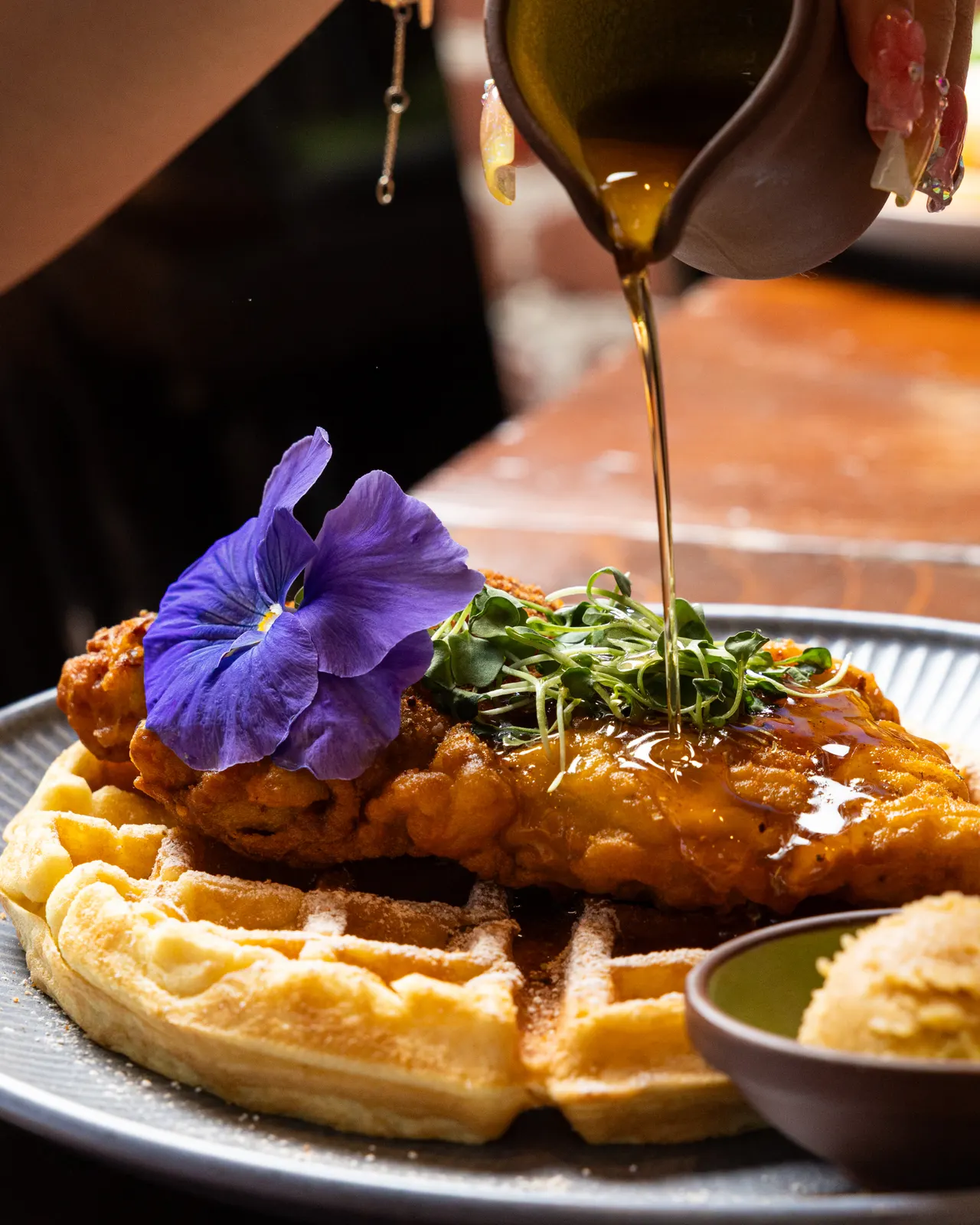 Syrup poured from a small ceramic pitcher over fried chicken on a waffle, a purple pansy and microgreens on top, at La Marcha, Berkeley