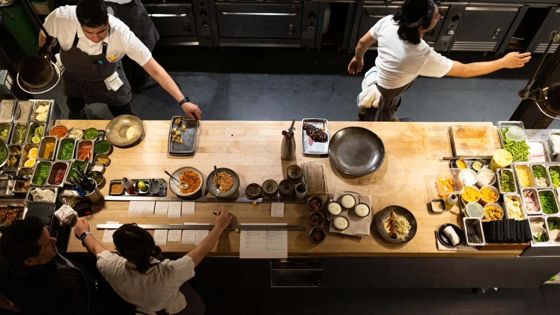 The kitchen pass from above at the OpenTable Ambassador Summit: cooks reaching across the butcher block, tickets on the rail and mise en place down both sides