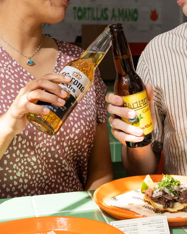Two guests clinking bottles over birria tacos under the tortillas a mano sign at Parranga, Santana Row