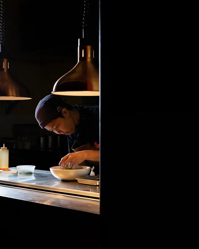 A line cook at Payakk, Oakland, plating under a copper pendant lamp in a dark kitchen