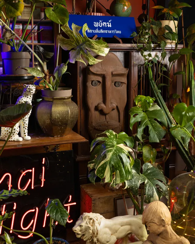 A carved wooden face, a brass urn and a ceramic leopard under the Thai sign, red neon glowing through the monstera at Payakk, Oakland