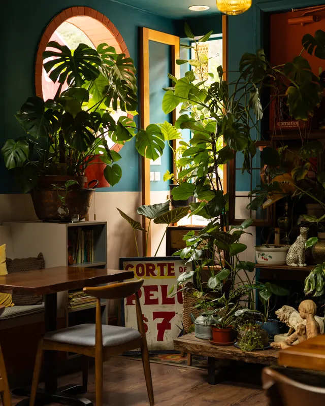 A corner table under monstera and a brick arch window, hand-painted signs and figurines on the shelves at Payakk, Oakland