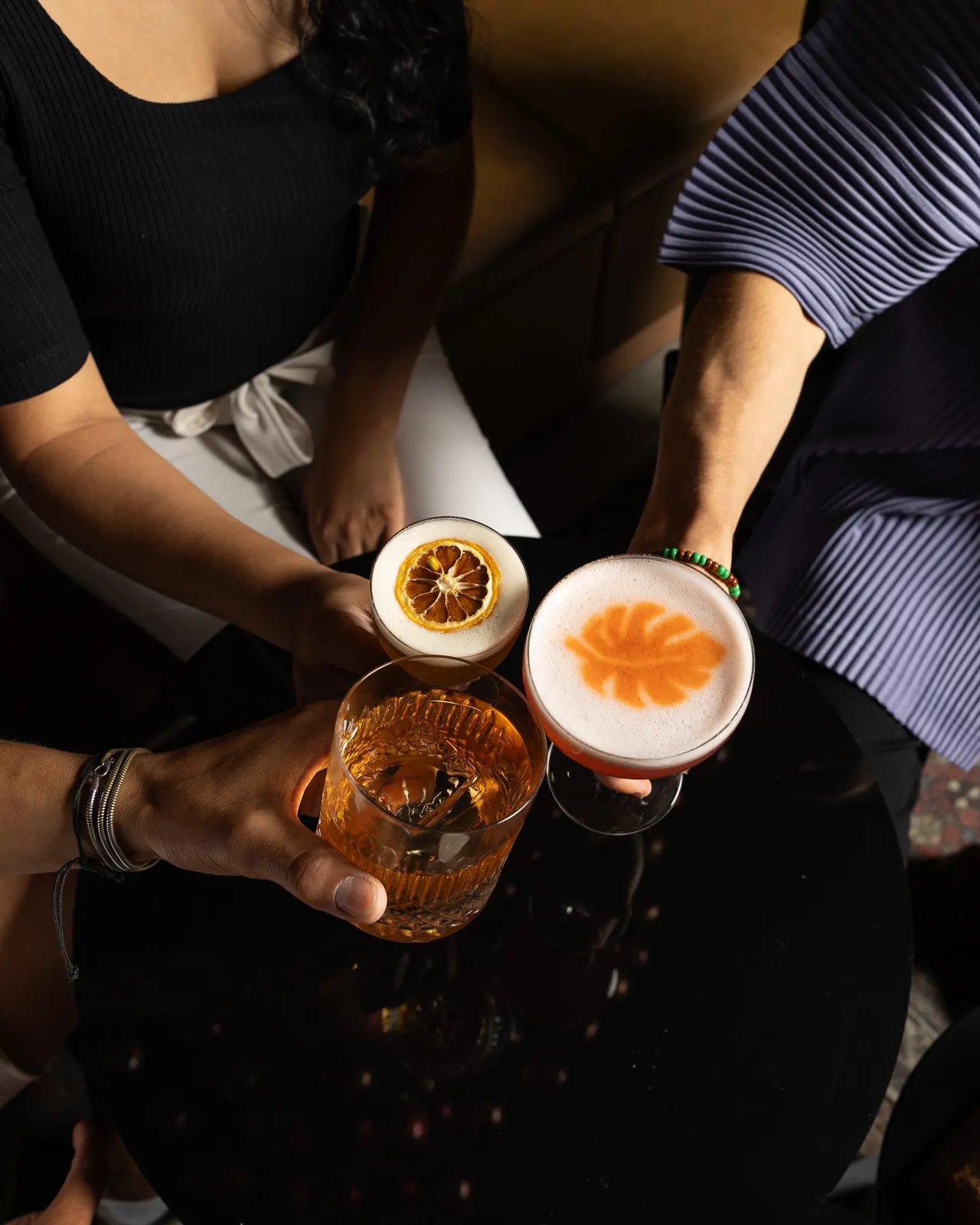 Three cocktails raised together from above, two friends leaning in over the black table at Suspiro, San Jose